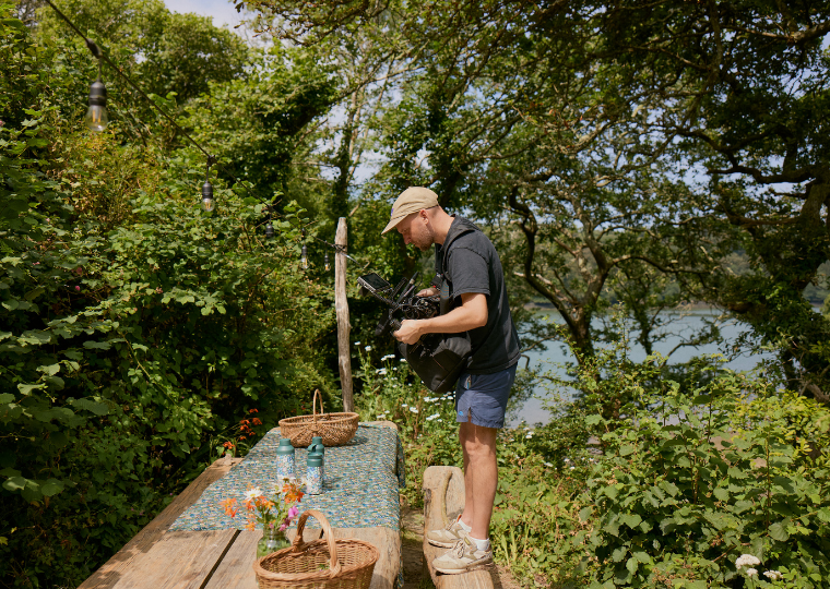 A man taking an aerial photograph of the Morley Blossom Ocean Bottle on a garden bench in a woodland area.